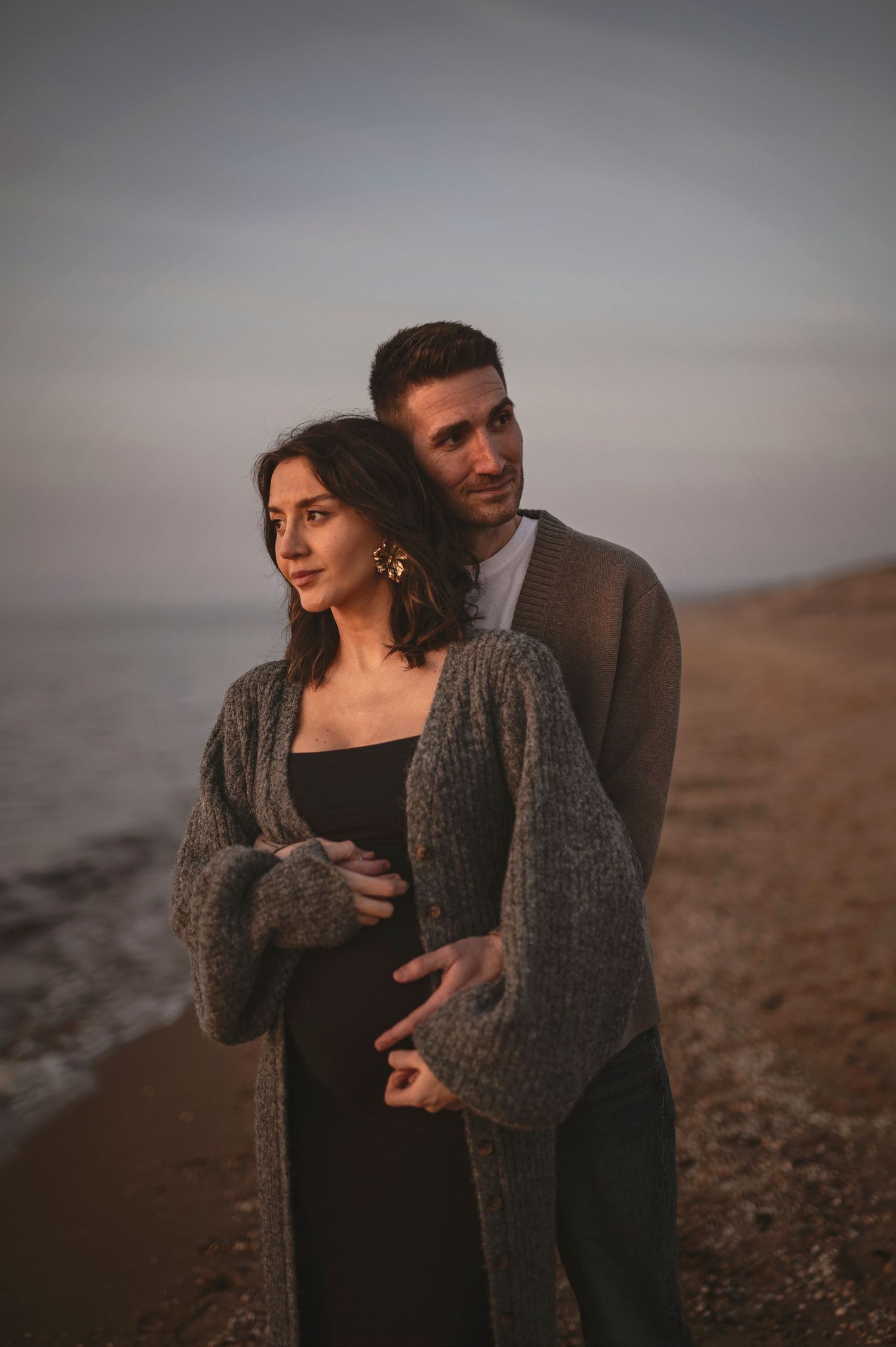 A romantic couple embraces on a moody beach at sunset, with the woman wearing a cozy gray cardigan over a black dress.