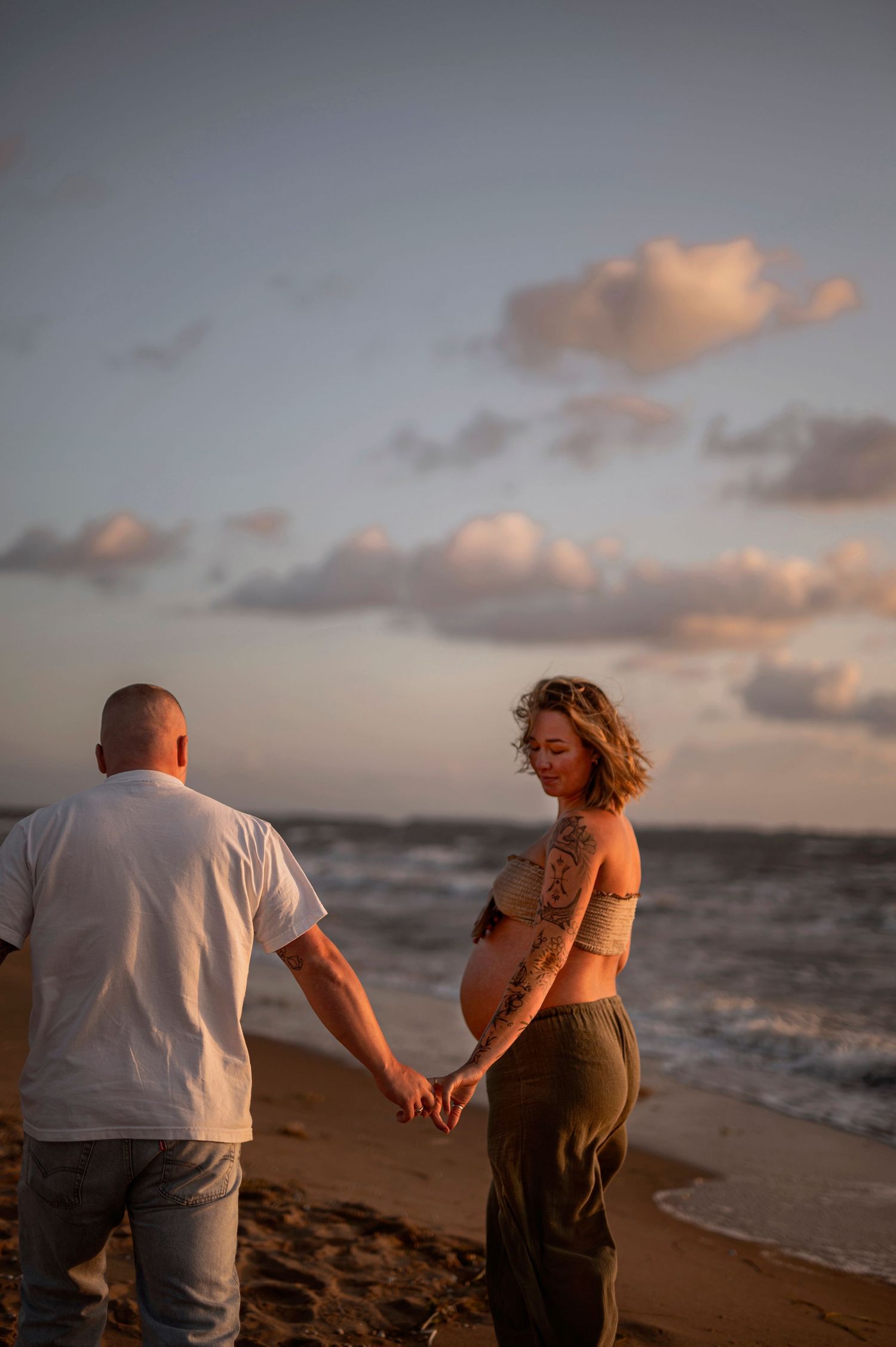 Couple holding hands on a sunset beach with waves crashing and golden clouds in the sky.