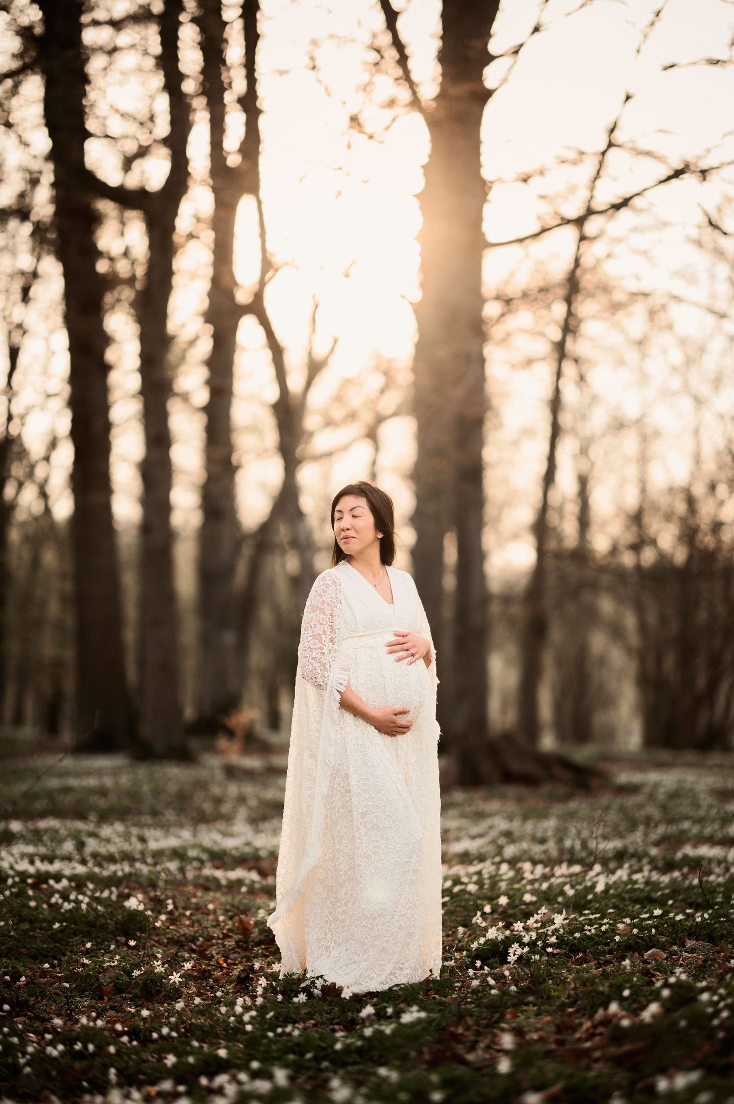Ethereal maternity photo series in a forest at sunset with white flowers blooming on the woodland floor.