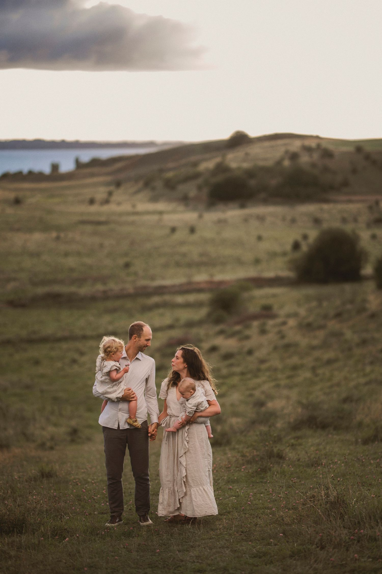 Family embraces together on a scenic hillside with water views in the background at golden hour.