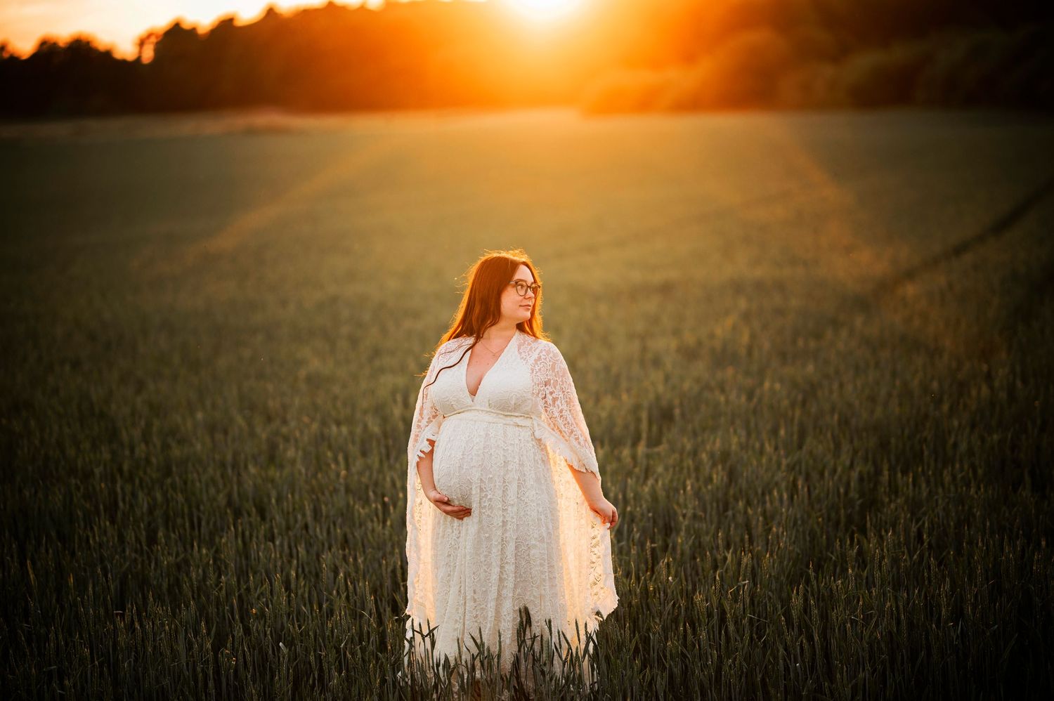 Dreamy maternity photo in white dress standing in golden wheat field at sunset.