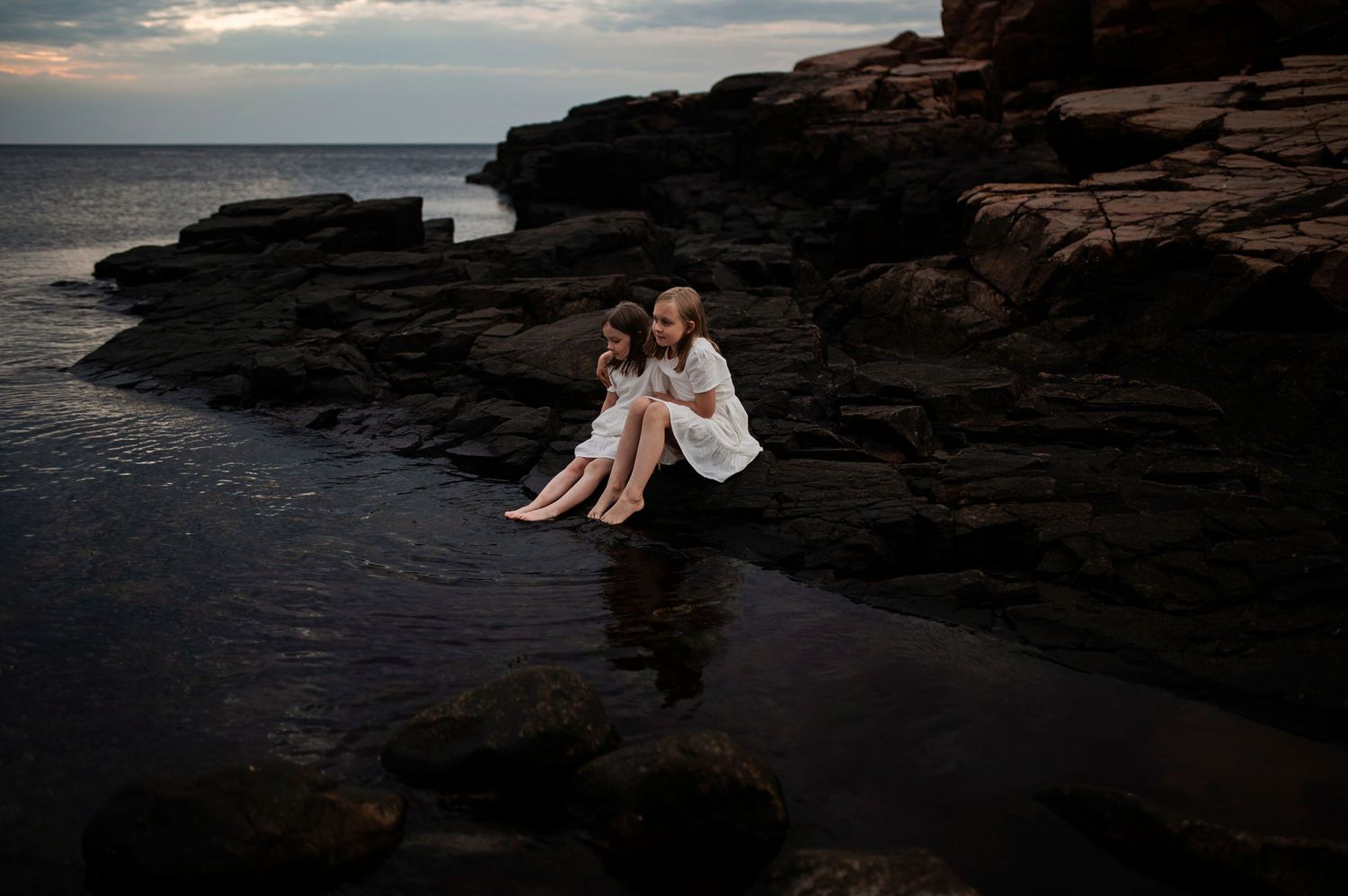 Two people in white dresses sit together on coastal rocks at dusk.