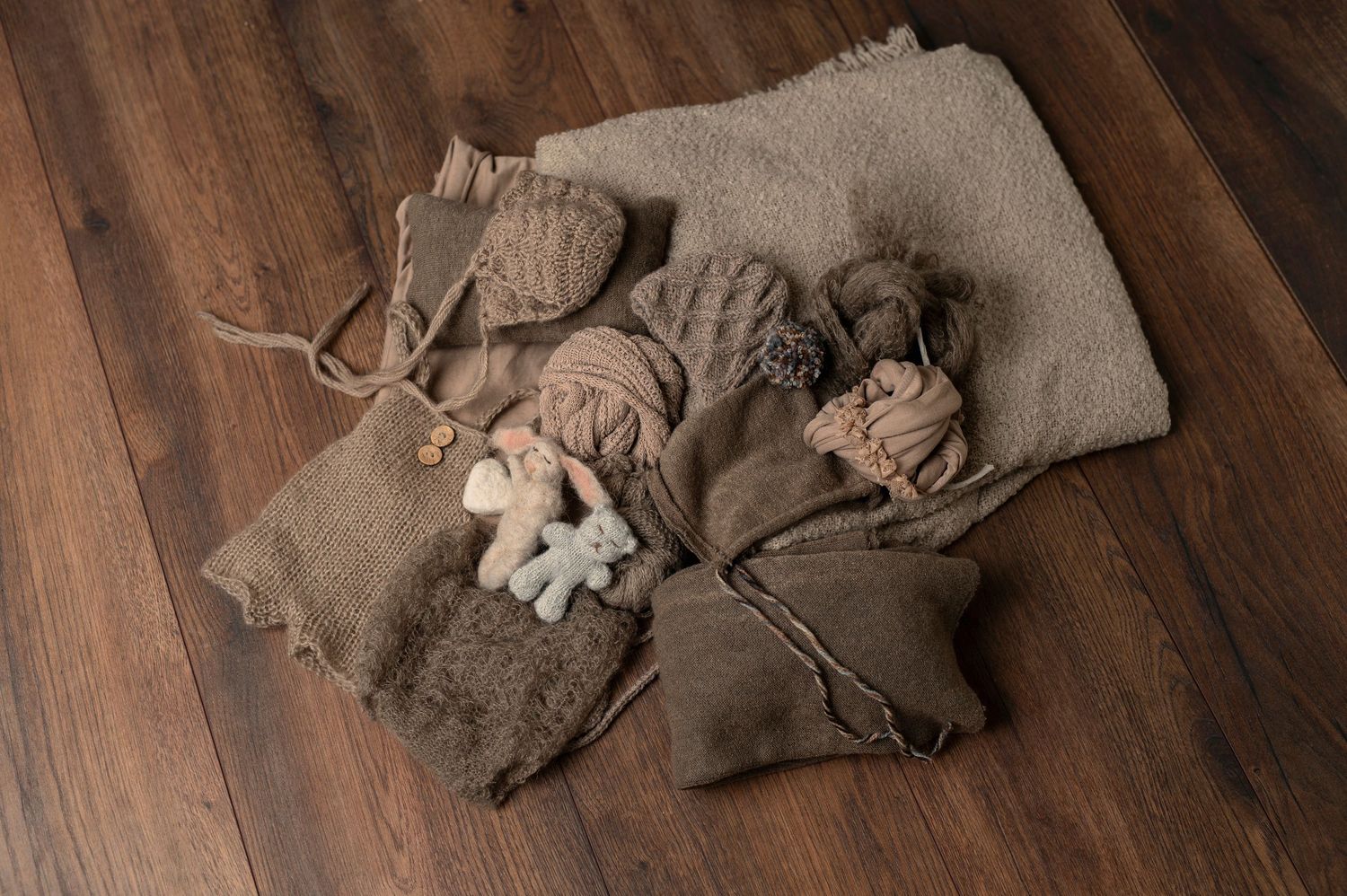 Neutral colored baby clothes and accessories arranged on dark wooden flooring.