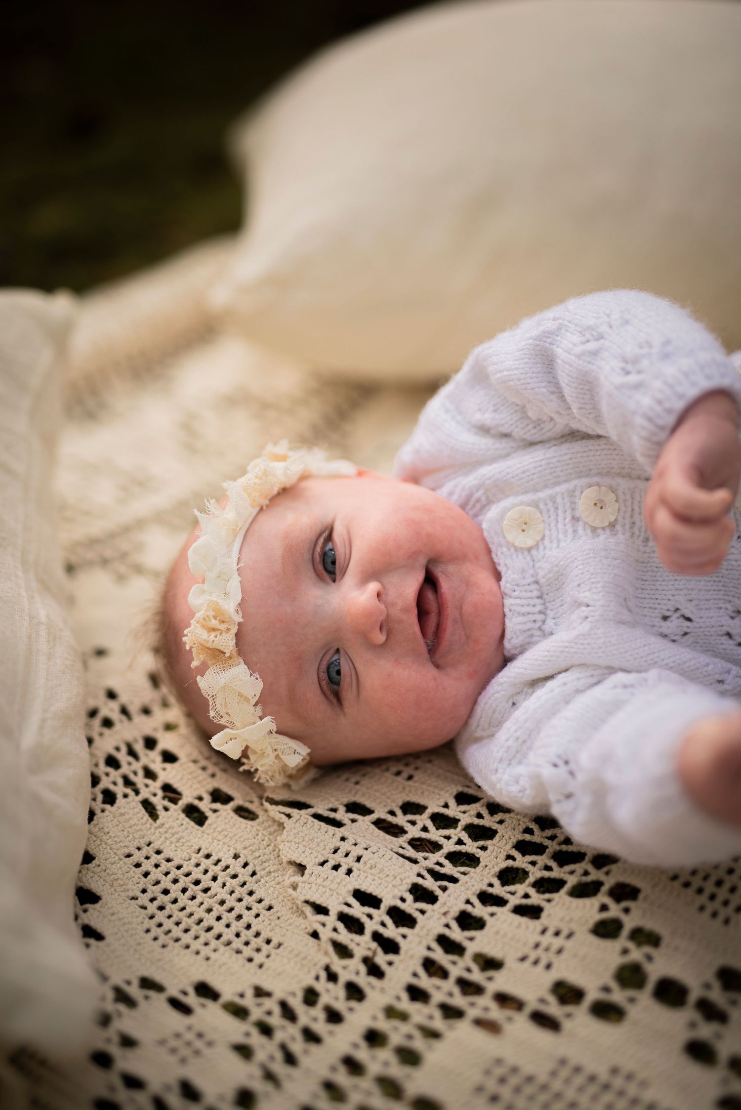 A baby in white lace clothing smiles while lying on vintage crochet blanket.