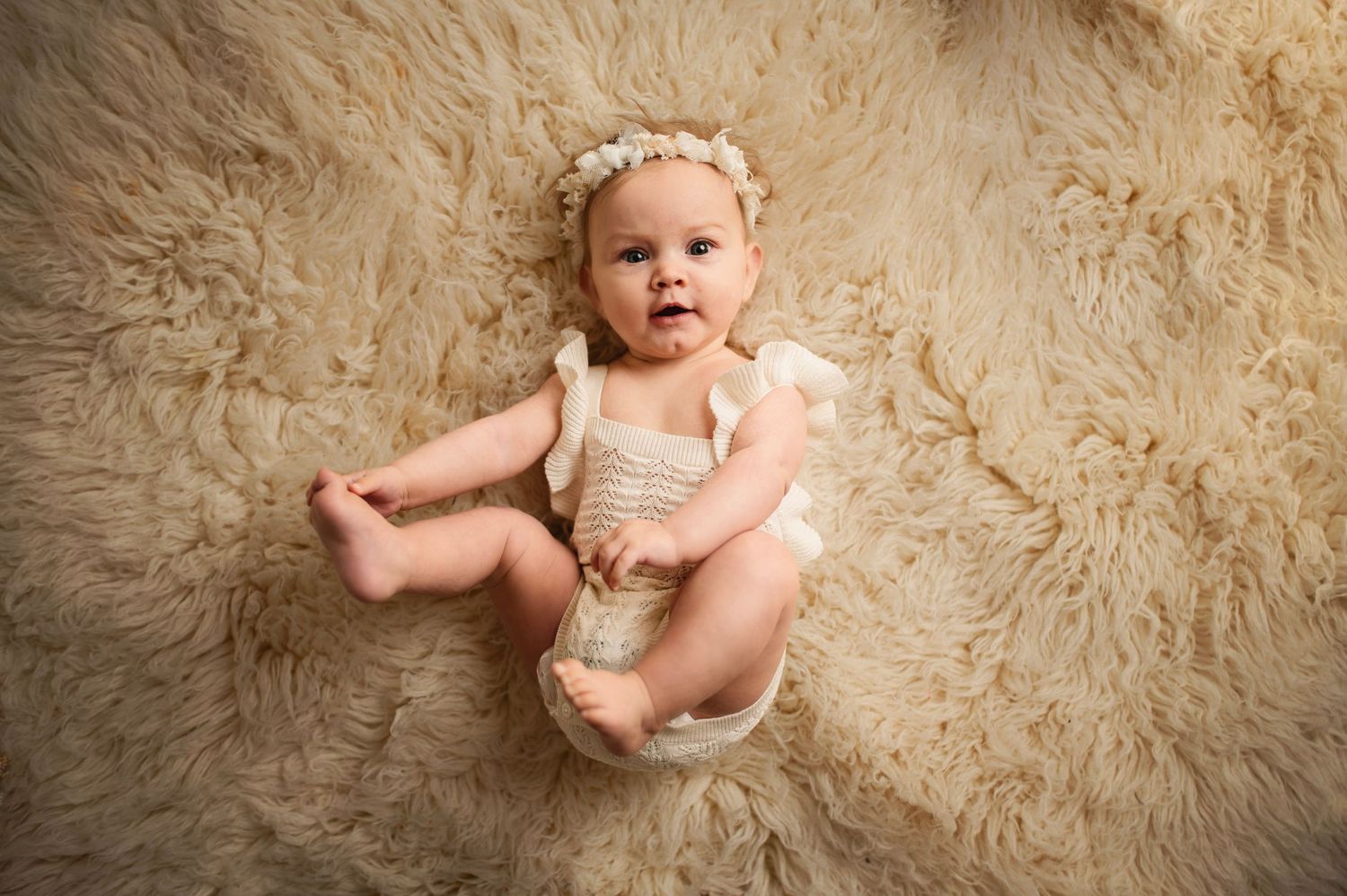 Infant in cream knit romper and flower crown posing on fluffy beige backdrop.