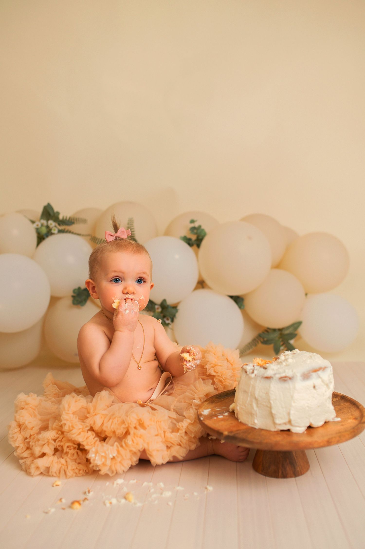 Baby sitting beside white cake on wooden stand with balloon backdrop during cake smash photography.