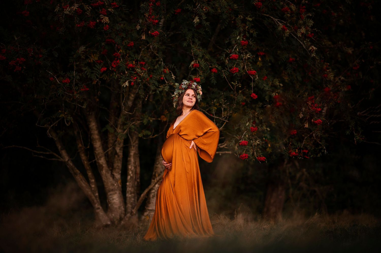 Pregnant woman in orange gown stands beneath red flowering tree wearing floral crown in dreamy outdoor maternity photo.