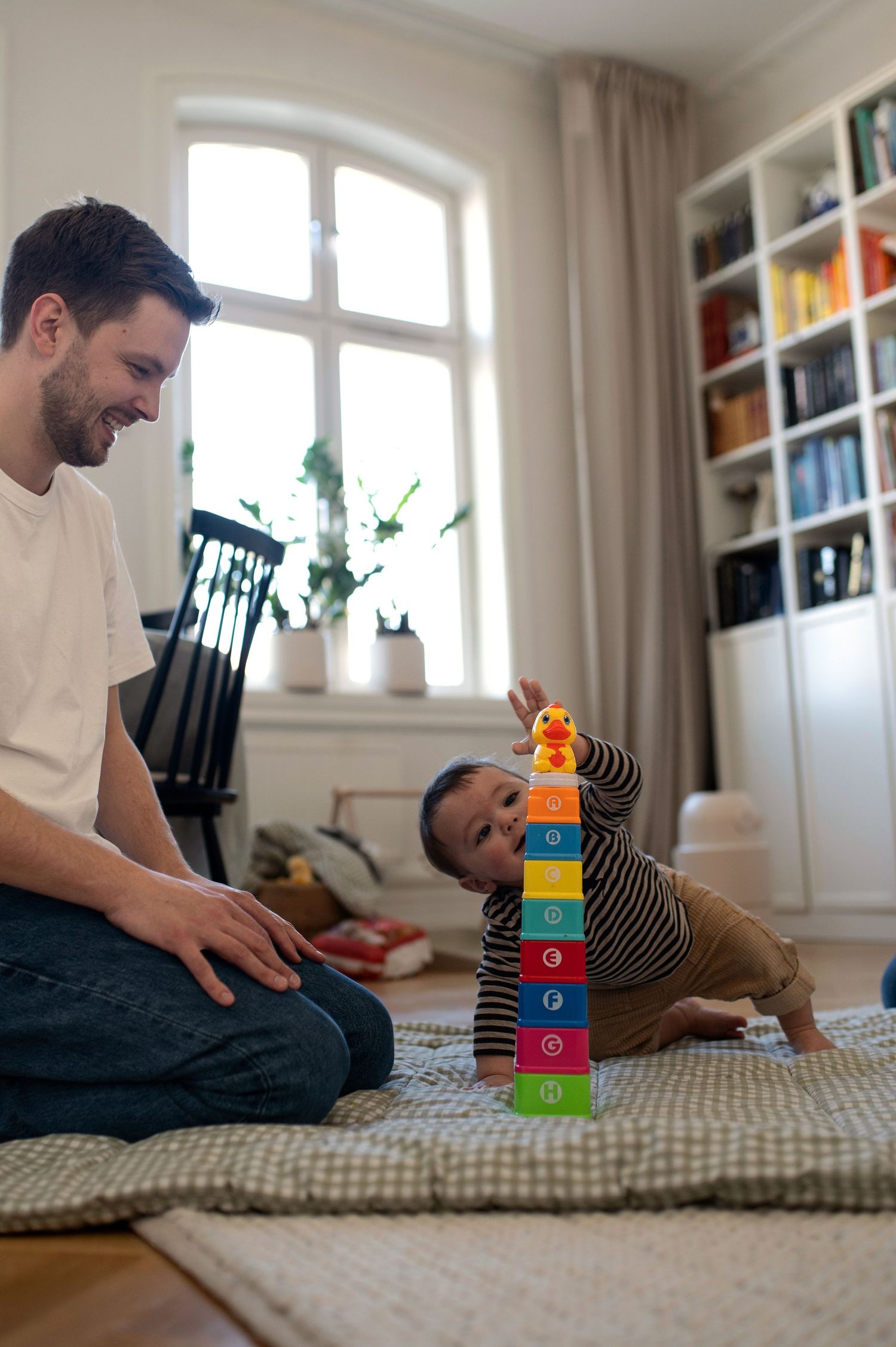 Happy father watching his baby stack colorful toy rings on the living room floor.