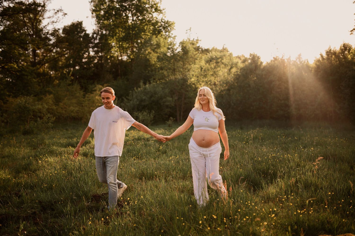 A couple holds hands while walking together through a grassy field at sunset with trees silhouetted in the distance.
