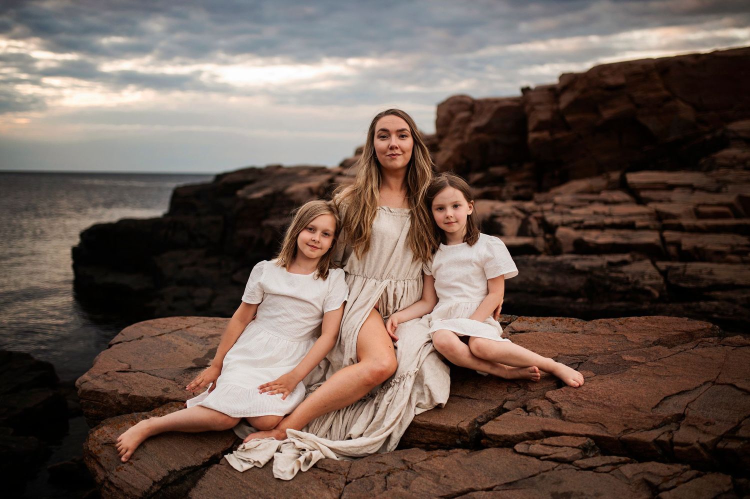 People in white and cream dresses pose on rocky coastal formation at sunset.