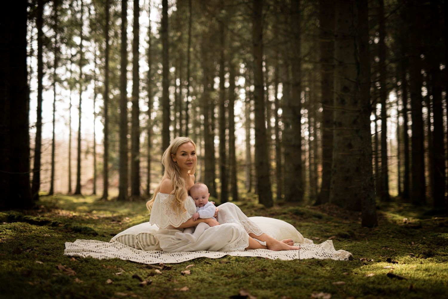 A dreamy forest maternity photo with sunlit trees and a lace blanket on mossy ground.