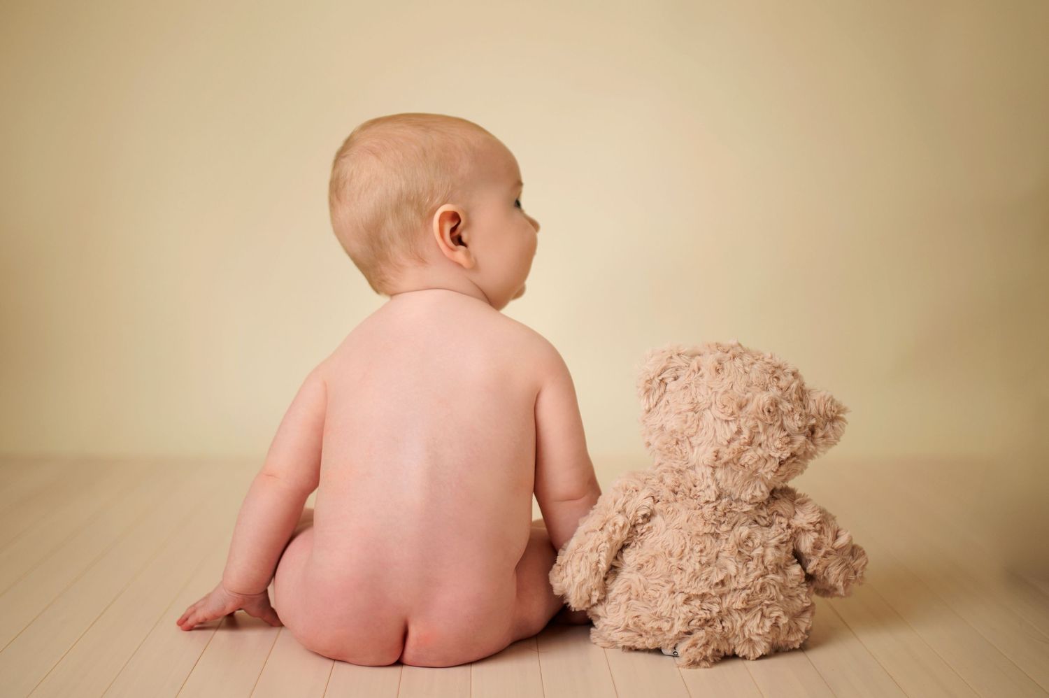 Back view of baby sitting next to plush teddy bear on beige background.