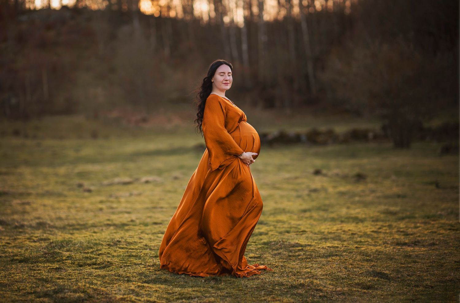 Pregnant woman in flowing burnt orange gown standing in sunlit green field during golden hour maternity photoshoot.