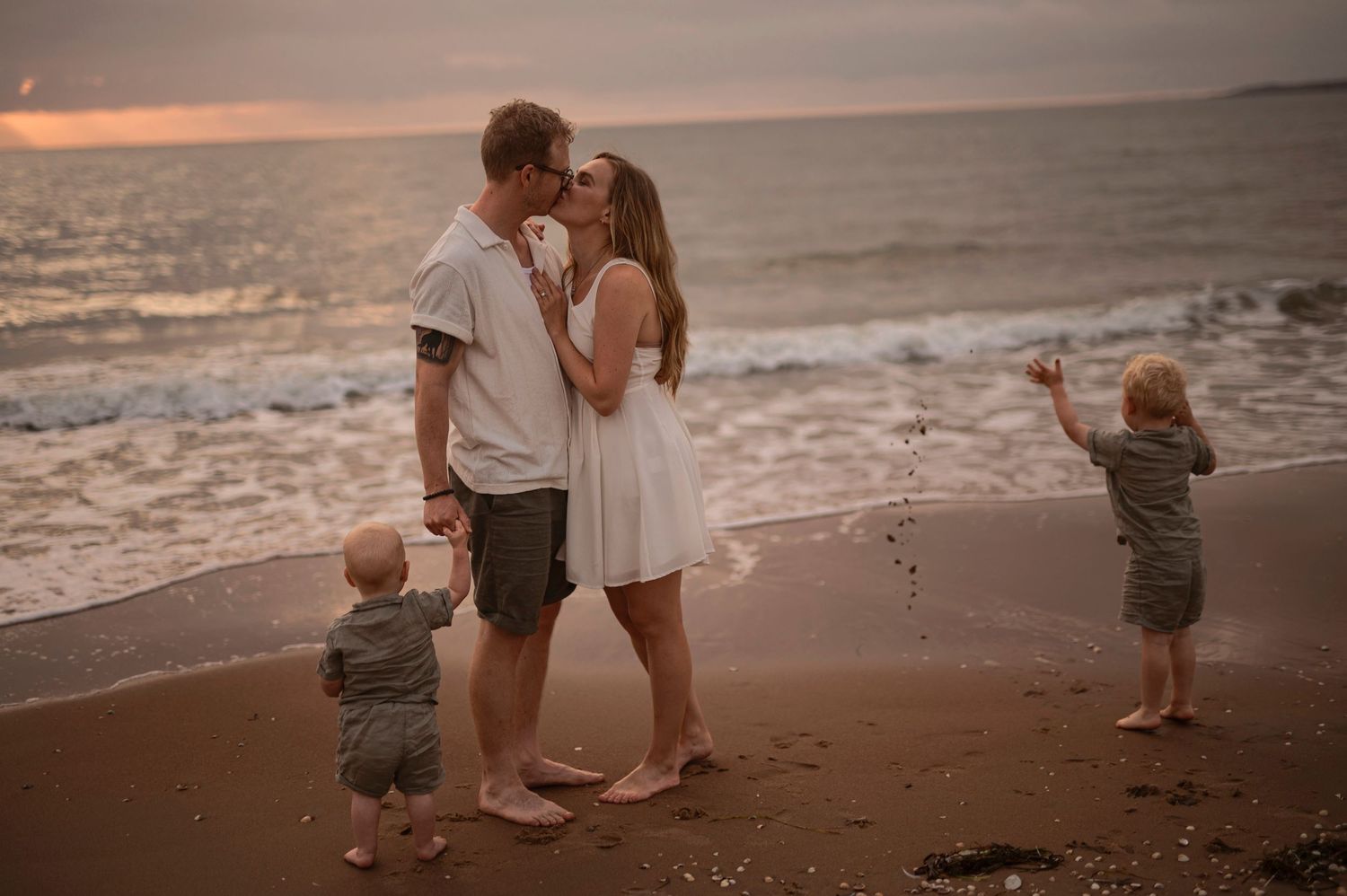 Family of four enjoying a romantic sunset beach moment, parents kissing while toddlers play on sandy shore.