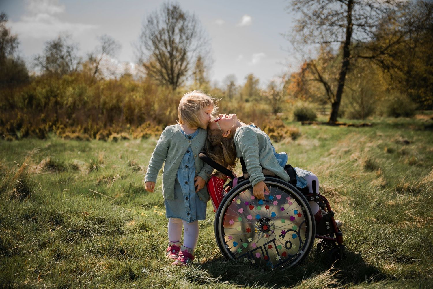 A young girl kisses a woman in a decorated wheelchair in a sunny green meadow surrounded by trees.