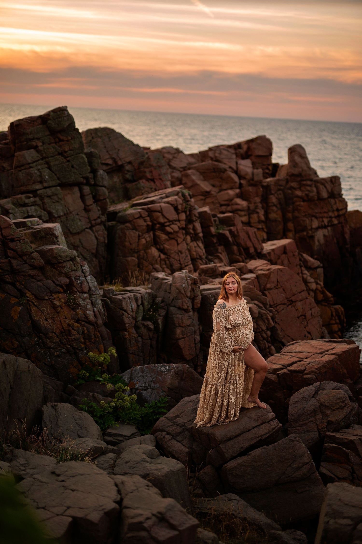 Woman in flowing dress poses on rugged coastal rocks at sunset.