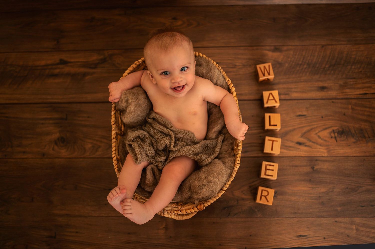 Baby resting in woven basket on wooden floor with letter blocks.