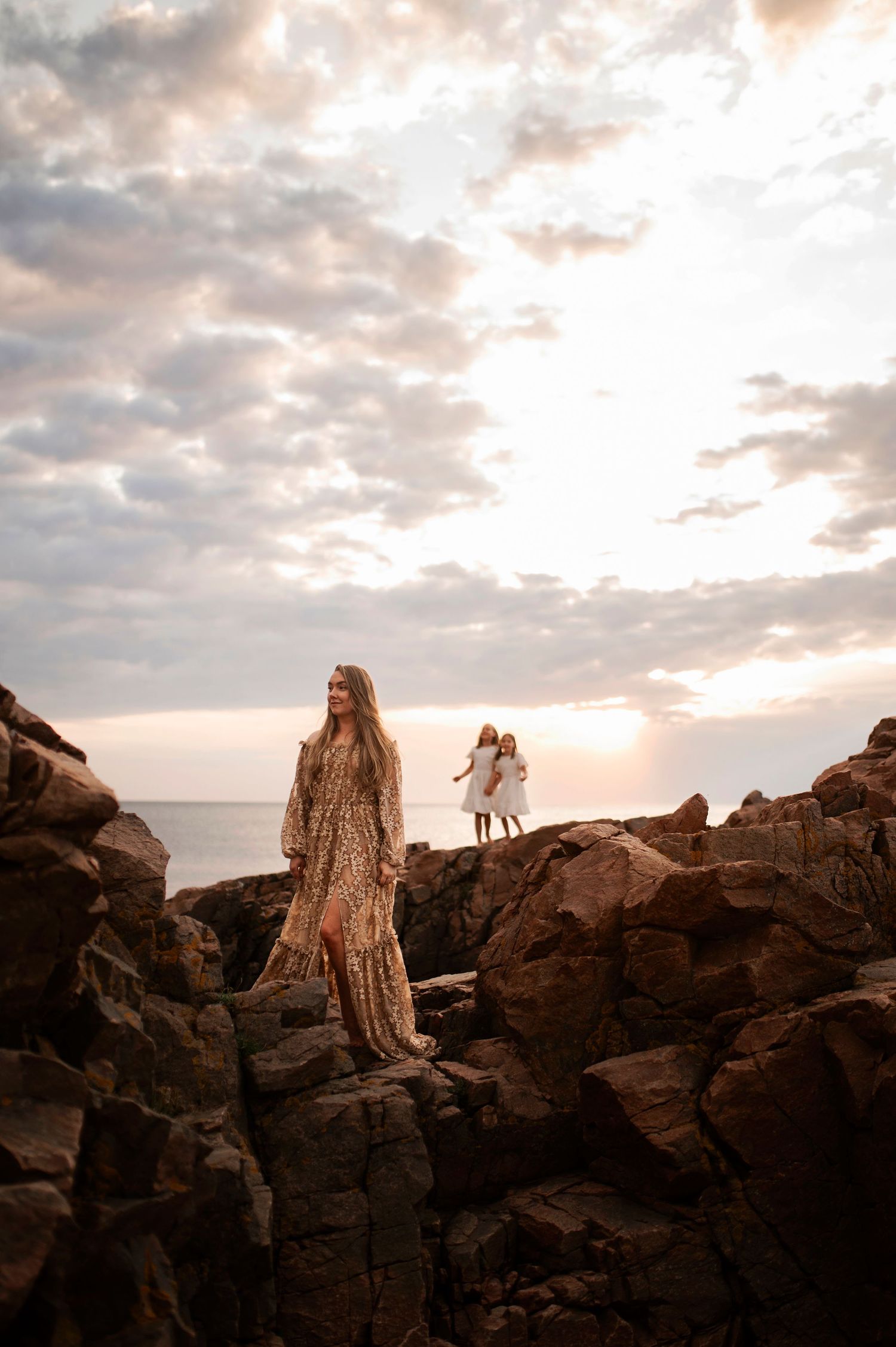 People in flowing dresses stand on dramatic coastal rocks during golden hour.