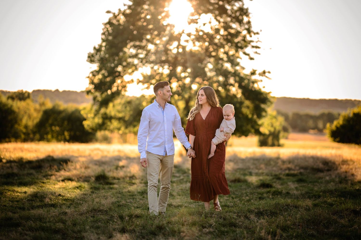A couple walking through a golden field at sunset with backlit trees and glowing warm light.