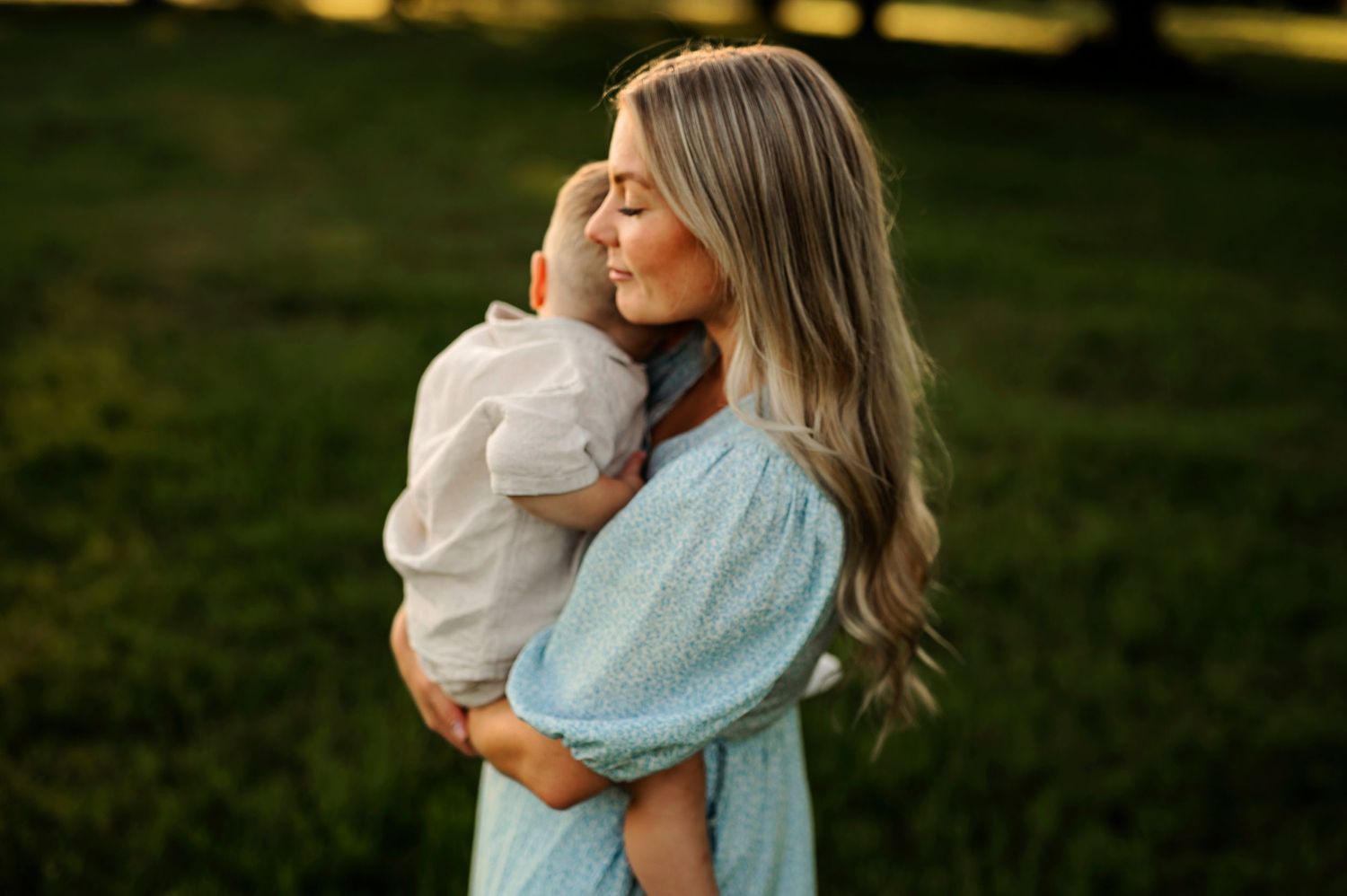 A tender moment captured at sunset as a light blue dressed figure cradles a small child in a natural outdoor setting.