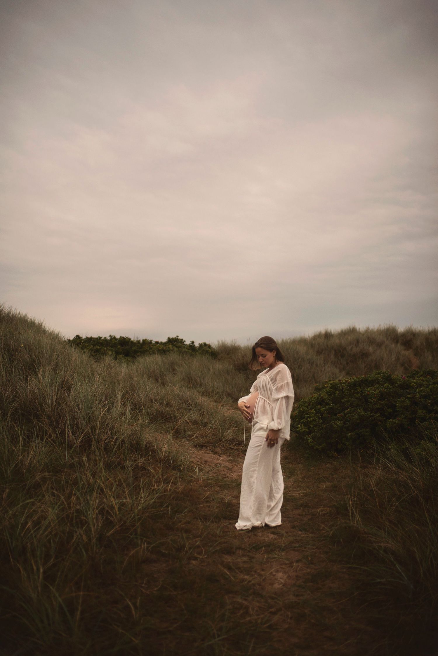 Person in flowing white dress poses on hillside path surrounded by coastal vegetation.