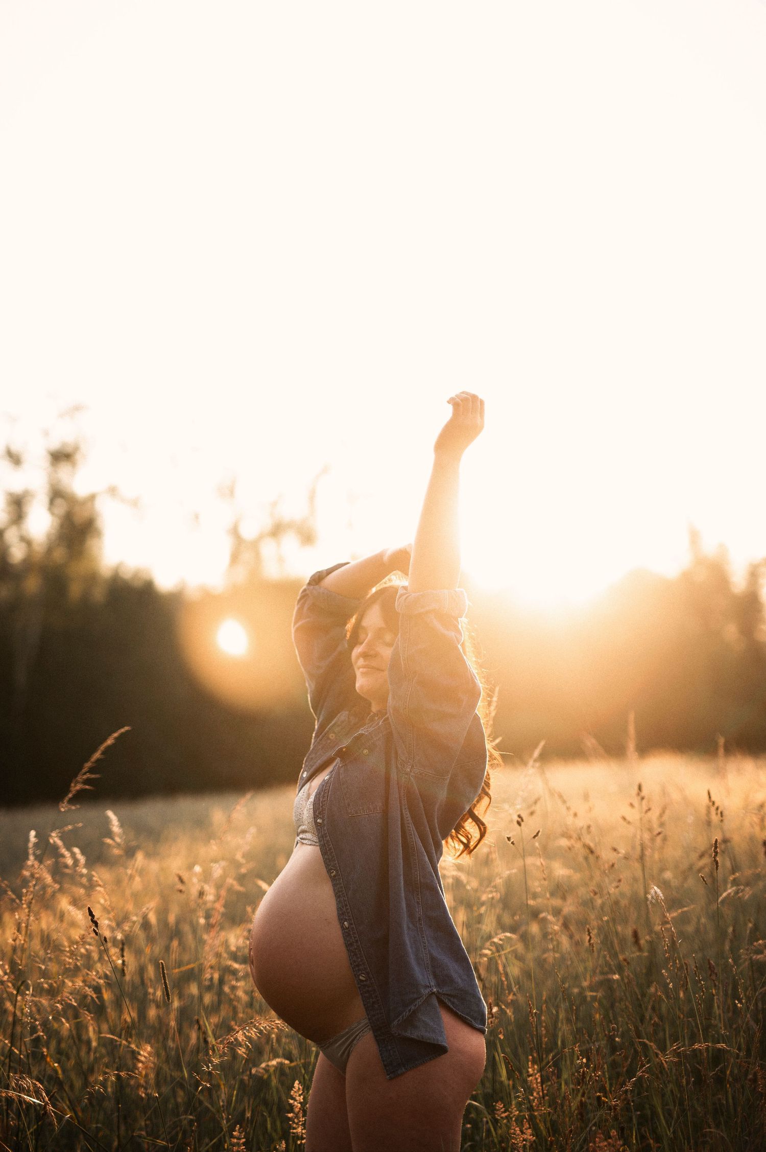 An expectant mother raises her arms victoriously in a field backlit by the setting sun.