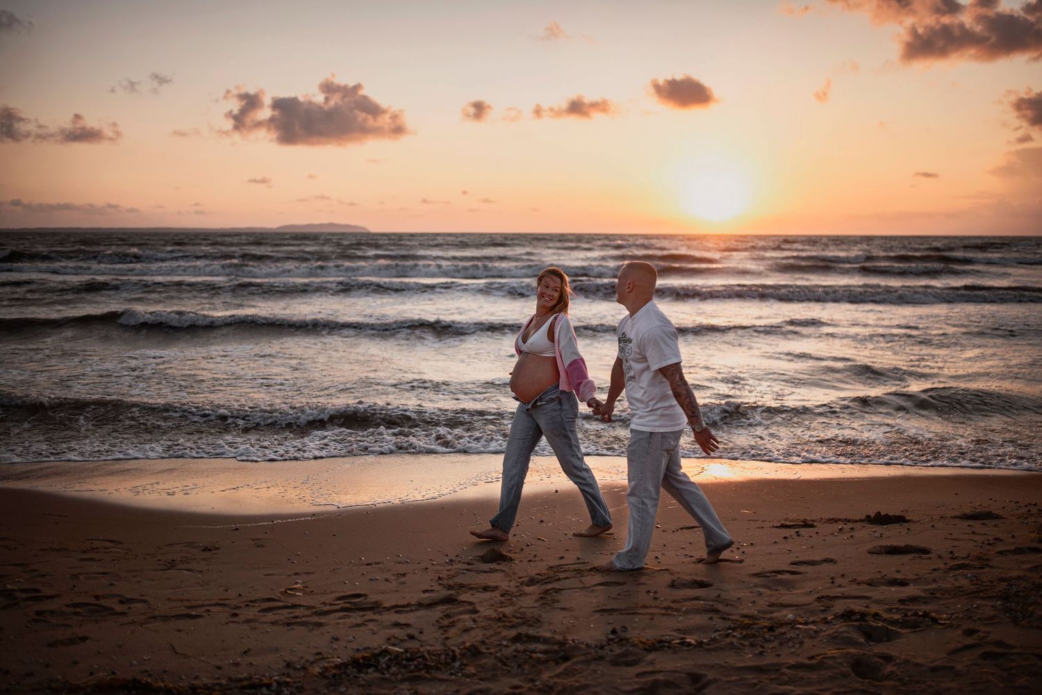 A couple walks hand in hand along a sandy beach at sunset with waves crashing in the background.
