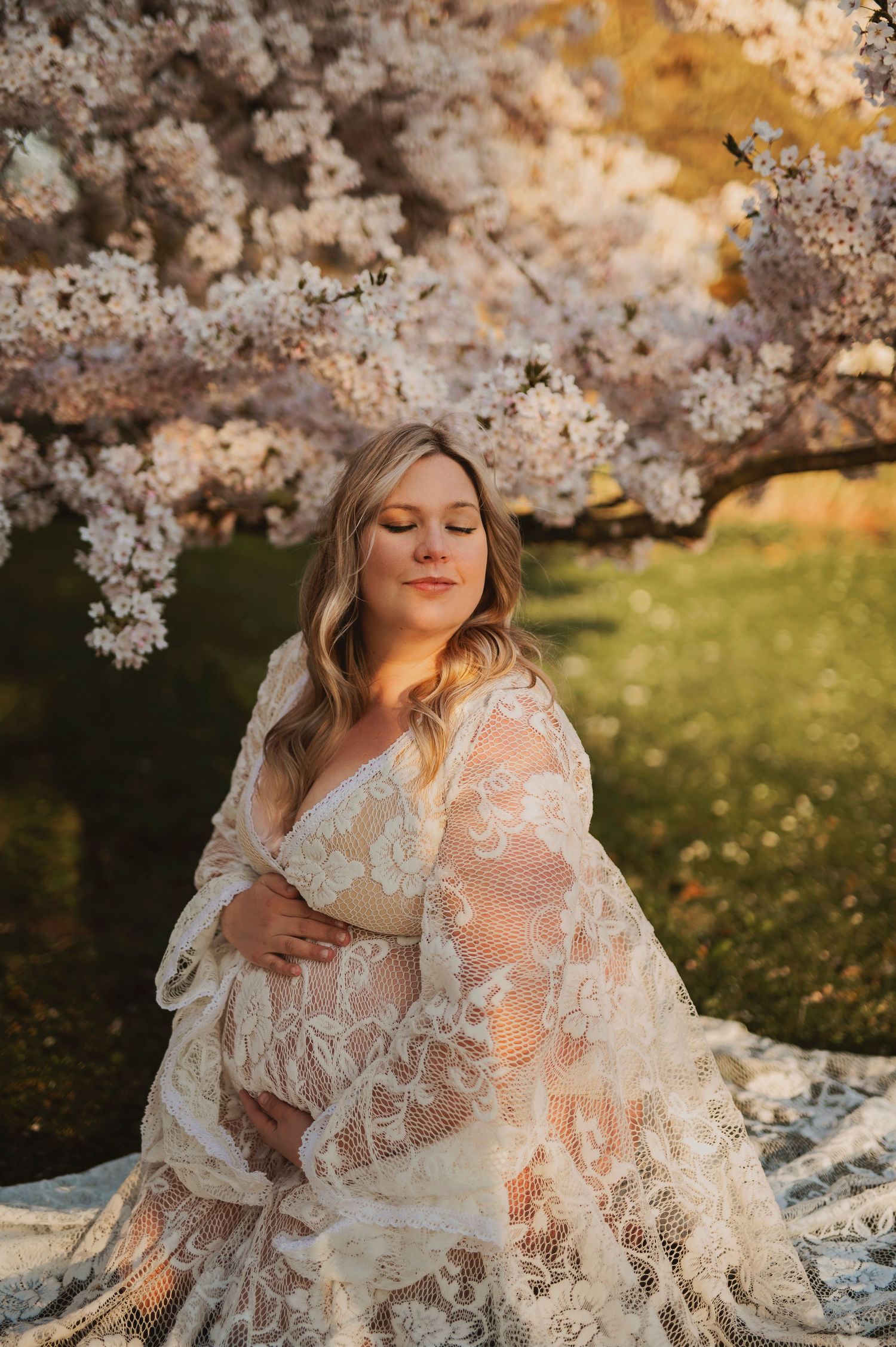 Elegant maternity portrait featuring white lace dress amid spring cherry blossoms.
