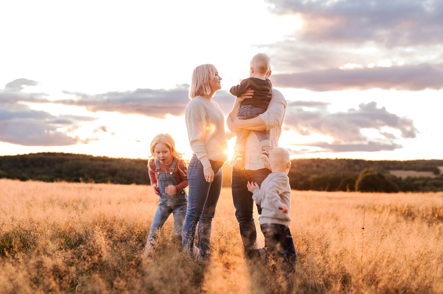 Family embracing in sunlit field with dramatic sunset sky.