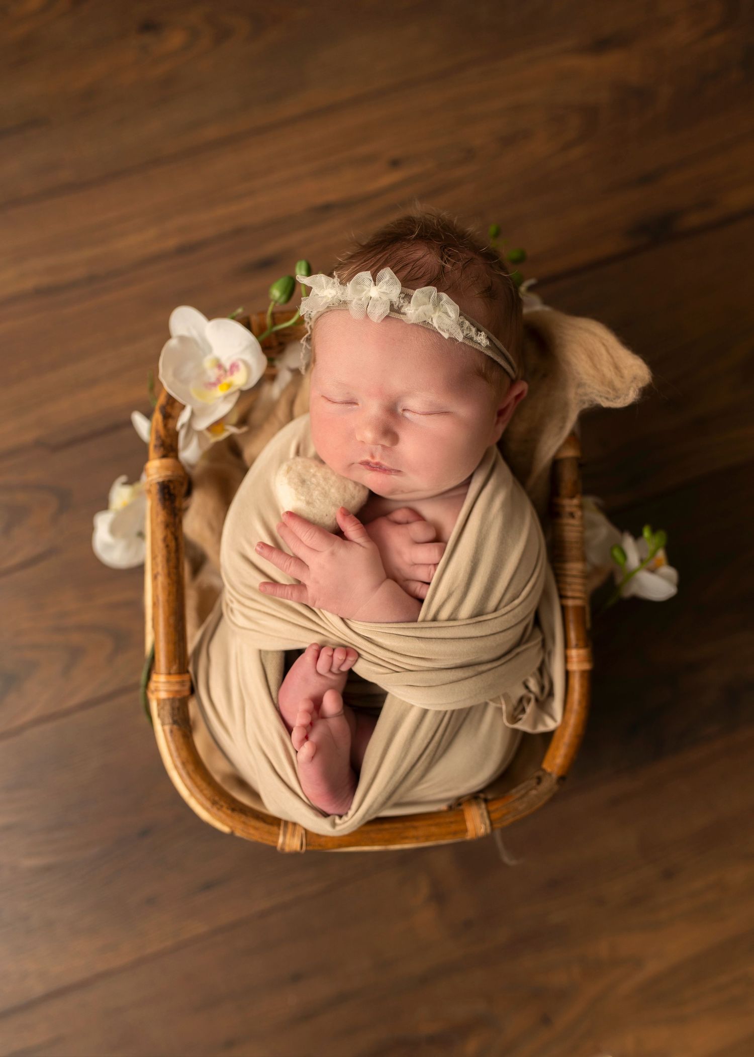 Sleeping newborn wrapped in beige fabric rests in woven basket with delicate floral decorations.