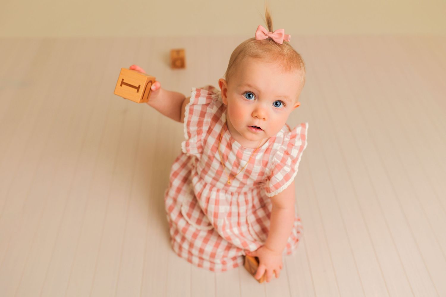 Baby in pink gingham dress sitting on cream backdrop during portrait session.