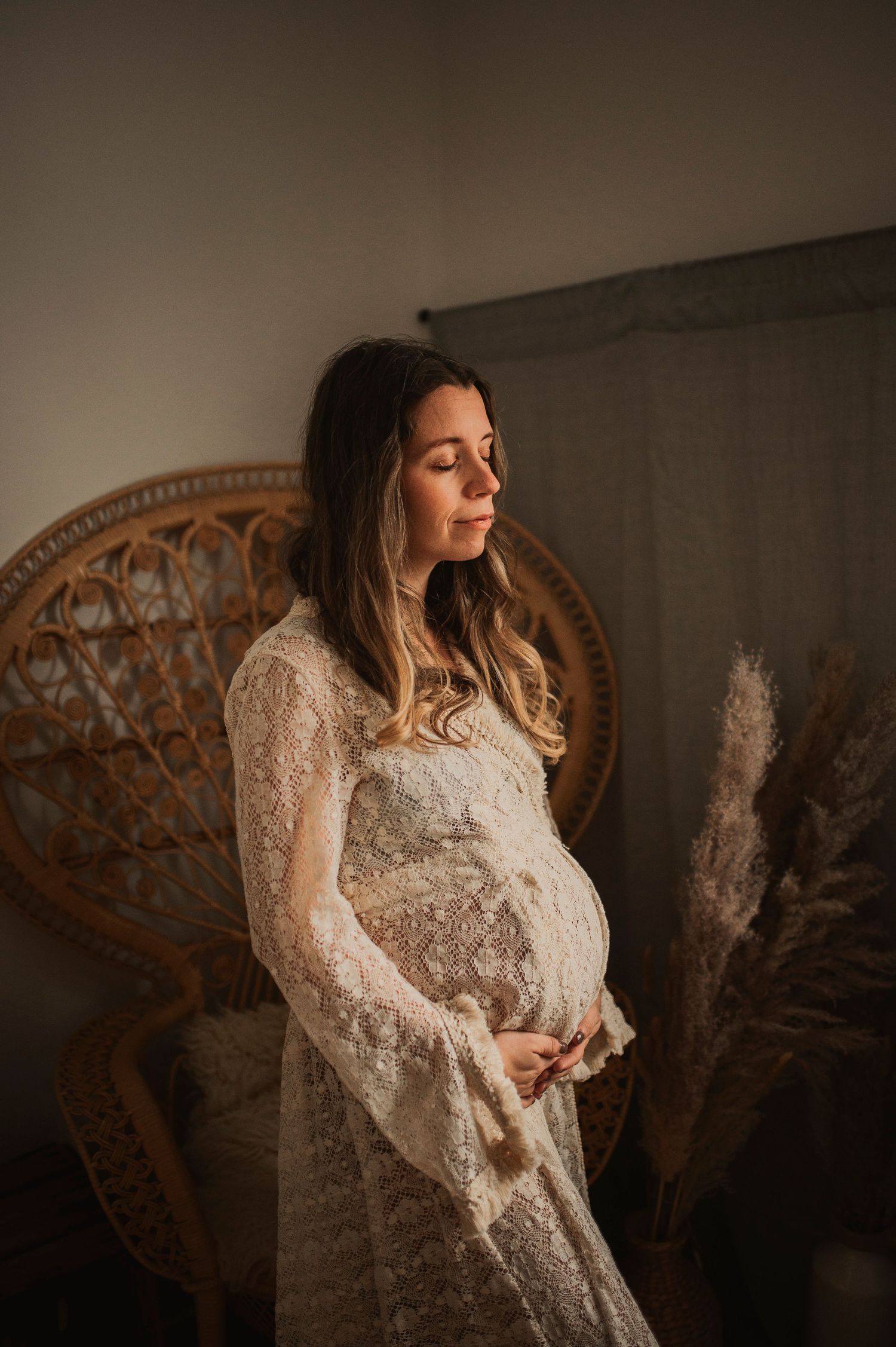 Ethereal maternity portrait of woman in lace dress beside woven peacock chair.