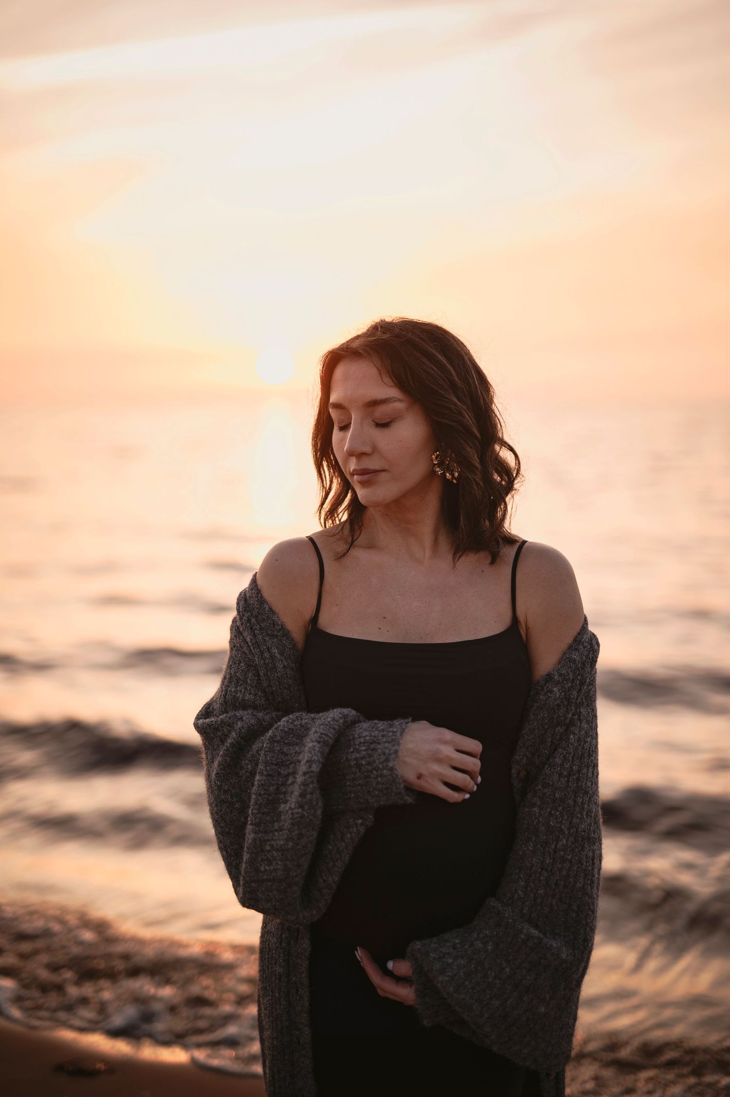 Pregnant woman in black dress and cardigan stands contemplatively by ocean at sunset.