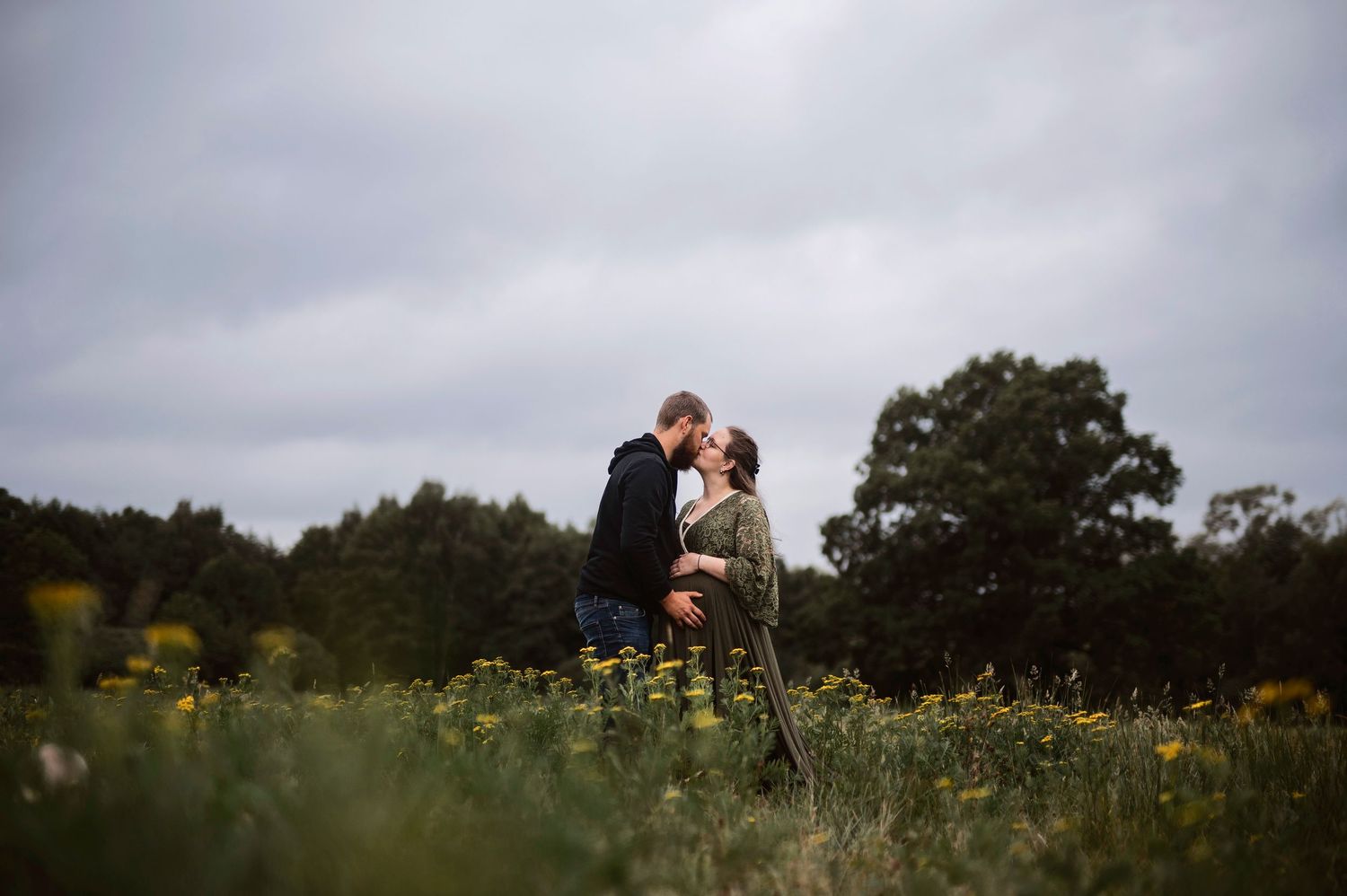 Couple sharing a romantic kiss in a wildflower meadow with lush green trees and dramatic cloudy sky backdrop.