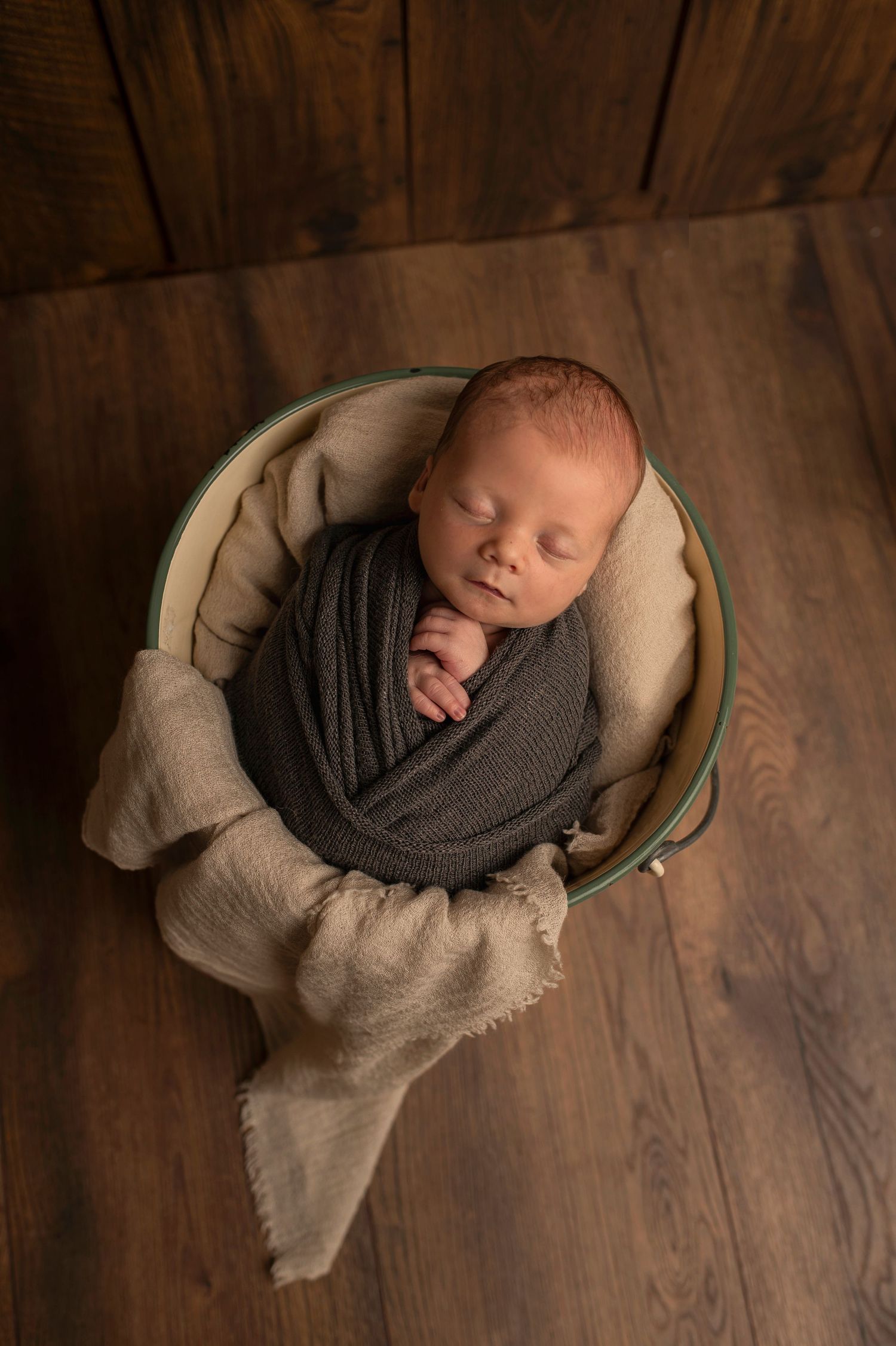Sleeping newborn wrapped in gray fabric nestled in a cozy cream-colored bowl on wooden floor.