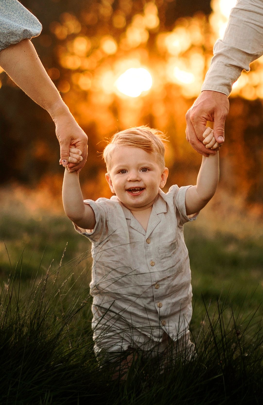 A small child in light clothing holds hands and smiles while walking through tall grass at sunset.