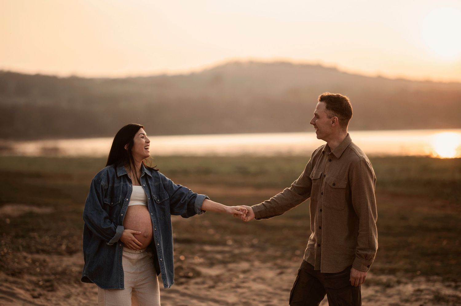 Couple stands together in warm sunset light on rocky terrain near water with mountains in background.