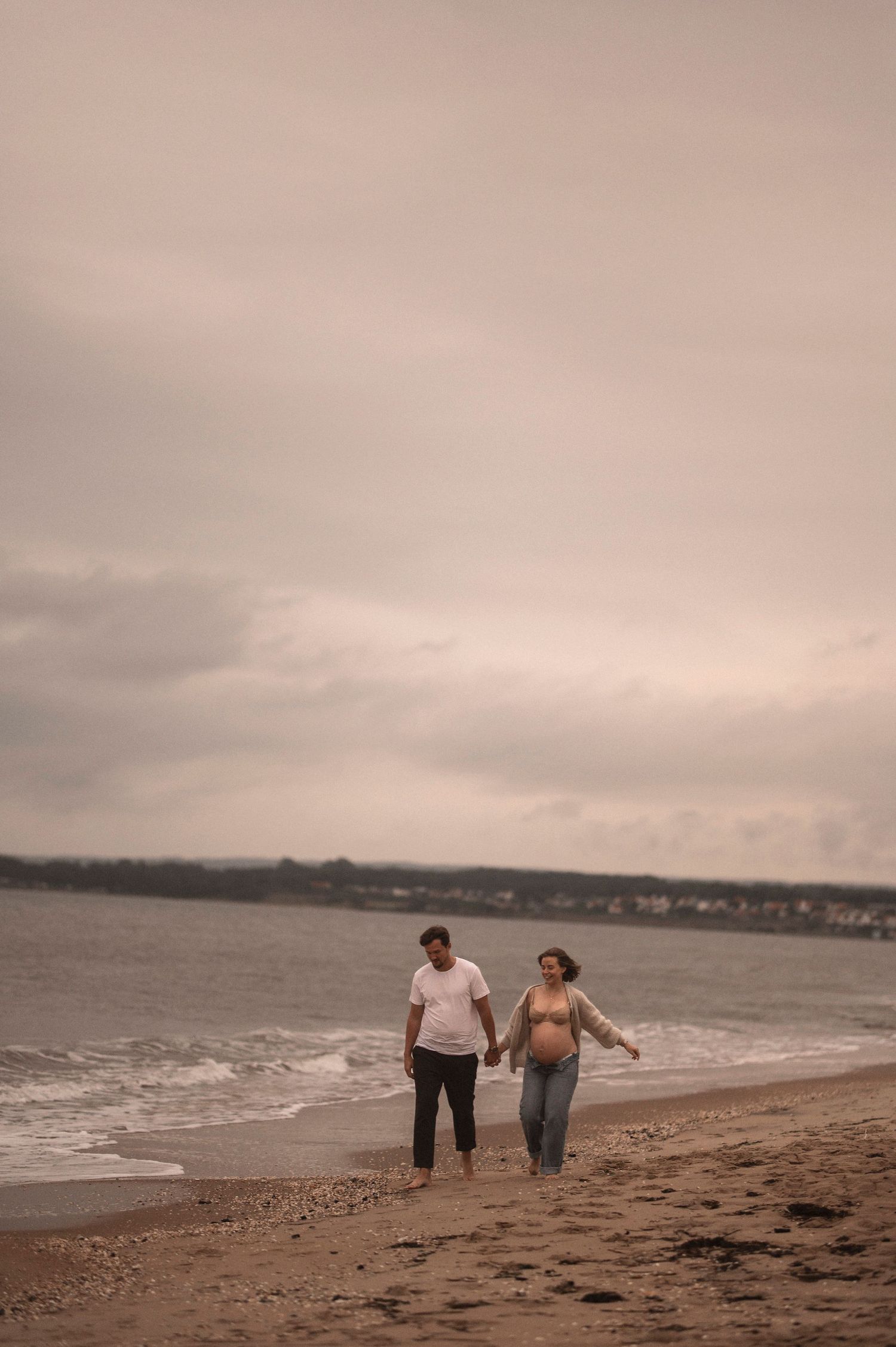 A couple walks hand in hand along a sandy beach with waves lapping at their feet.