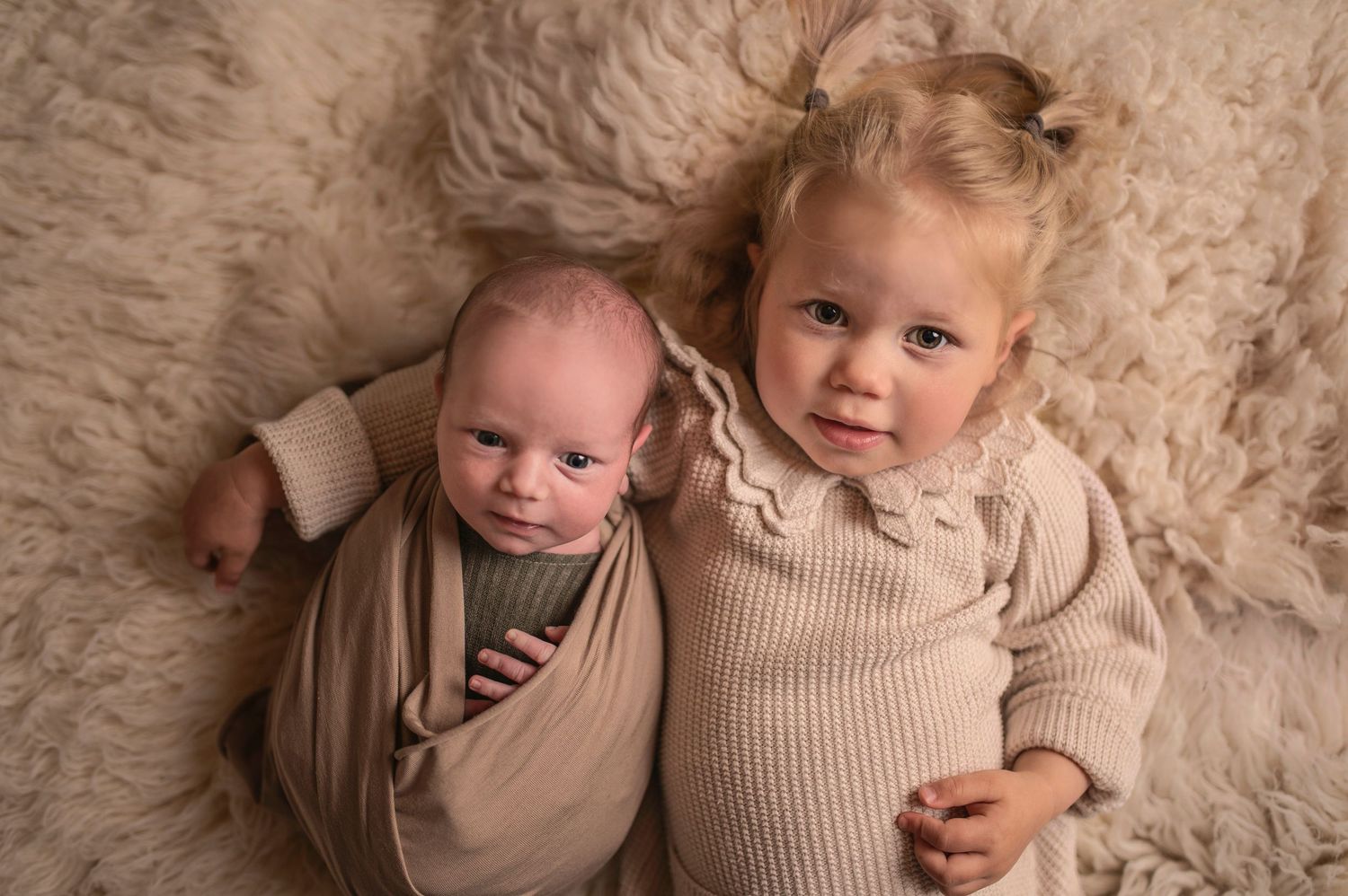Two young children in cozy beige sweaters lying together on a soft furry rug.