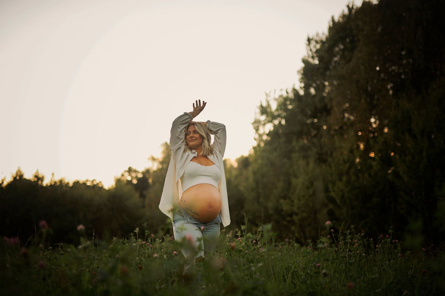 A maternity photo of an expectant mother in a field at sunset wearing a white top.