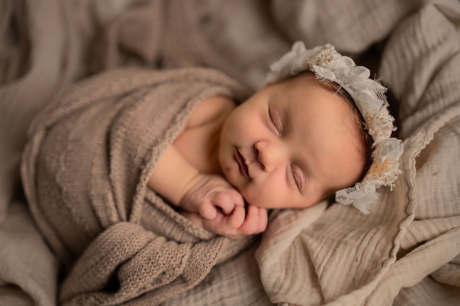 Peaceful sleeping newborn wearing a lace bonnet wrapped in soft beige knit fabric.