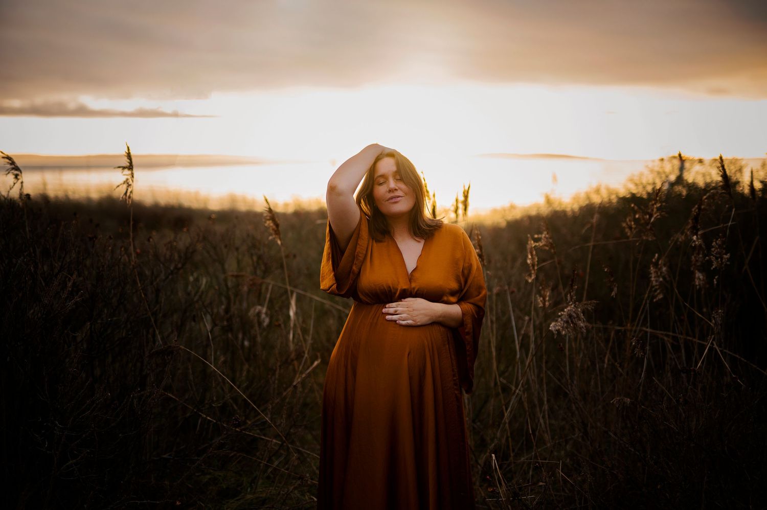 A figure in a rust-colored dress stands in a field backlit by golden sunset light.