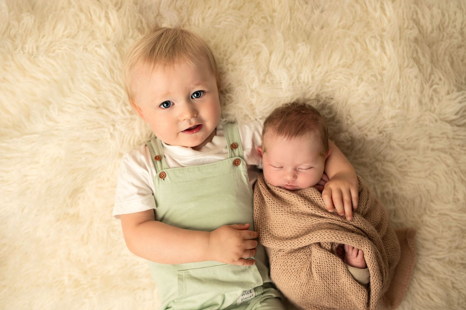 Two young children on soft white fur, one in green overalls and one wrapped in beige blanket.
