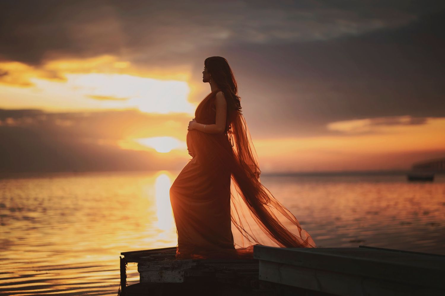 A silhouetted figure in flowing dress stands on dock during golden sunset over water.