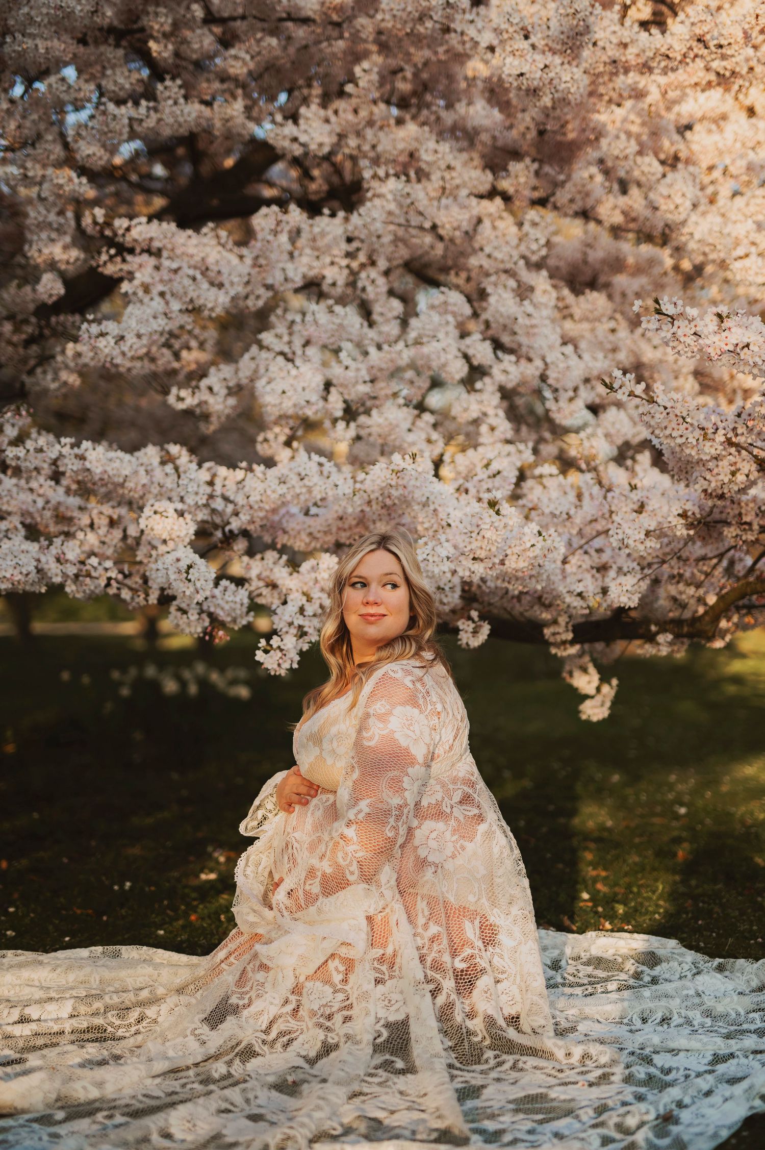 Artistic maternity photo with flowing lace gown under cherry blossoms at sunset.