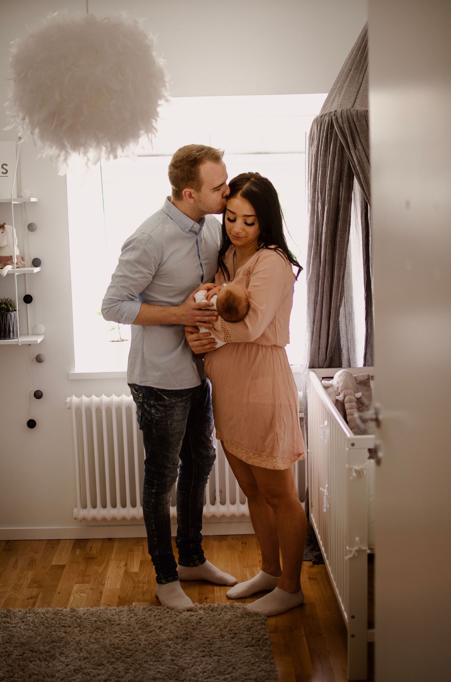 New parents sharing tender kiss while holding newborn baby in nursery room.