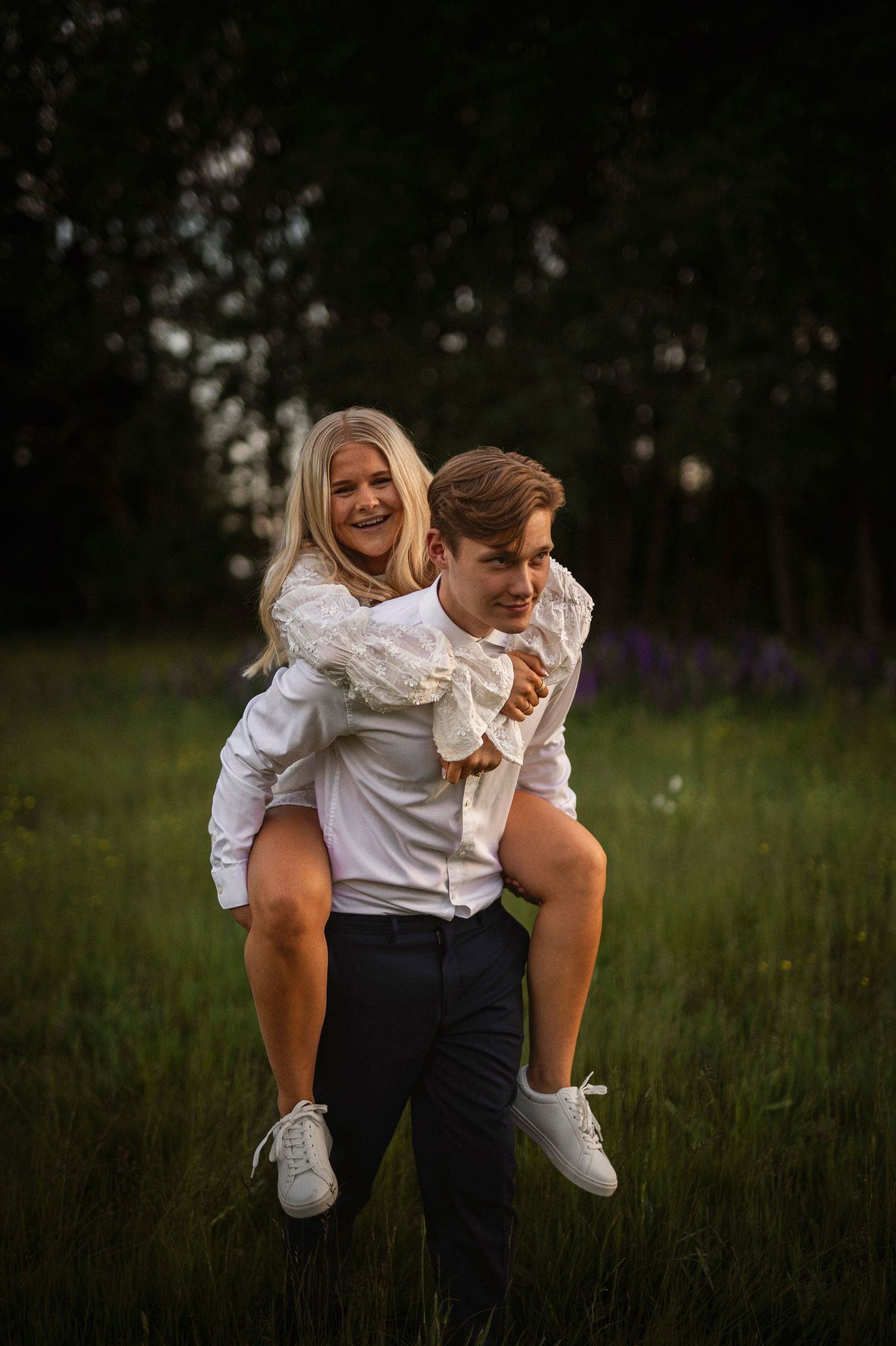 A young man gives a laughing blonde woman a piggyback ride through a sunlit green field at sunset.