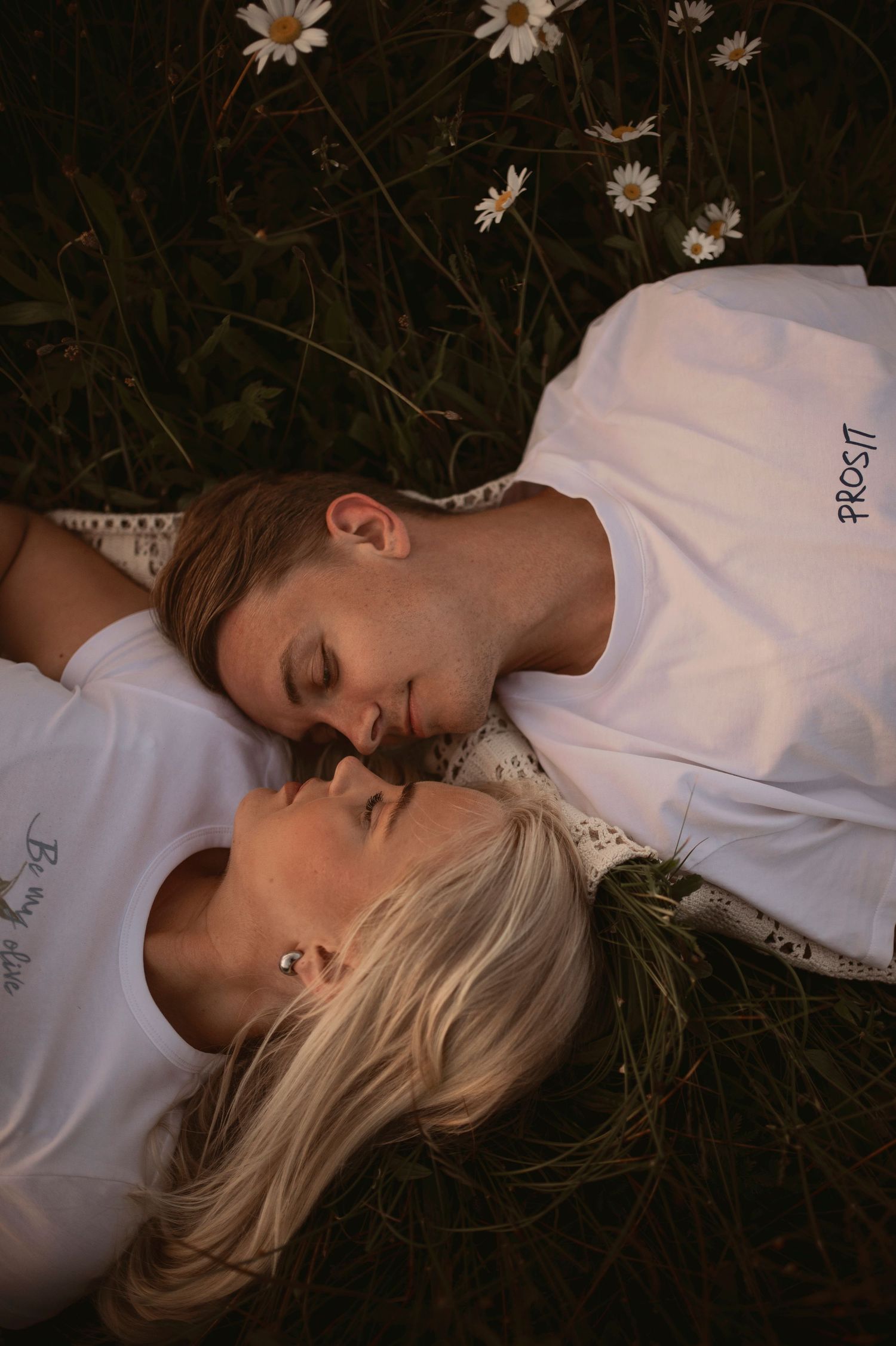 A couple lies together in a meadow with white daisies while wearing white shirts, photographed from above.
