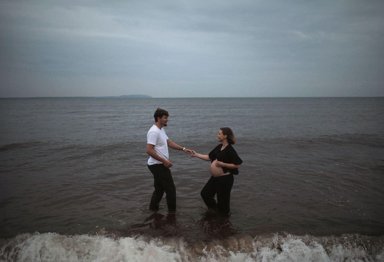 Expecting couple holding hands while standing in the ocean waves.