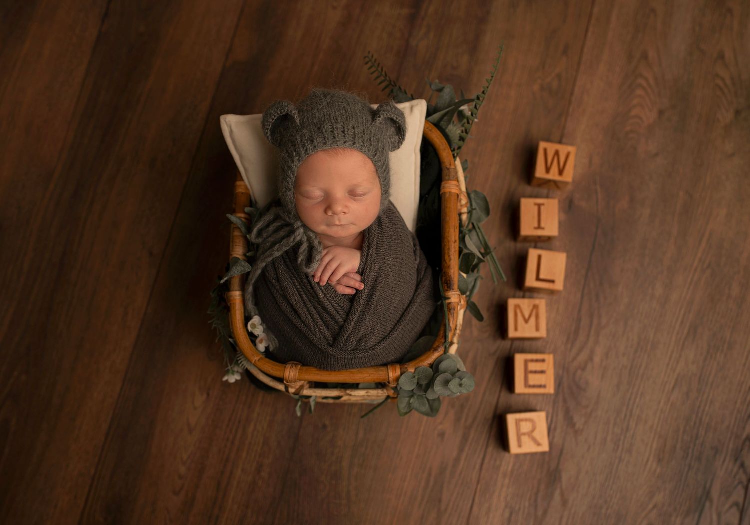 Newborn baby sleeping peacefully in a gray wrap inside a wooden basket with wooden letter blocks spelling WILDER.